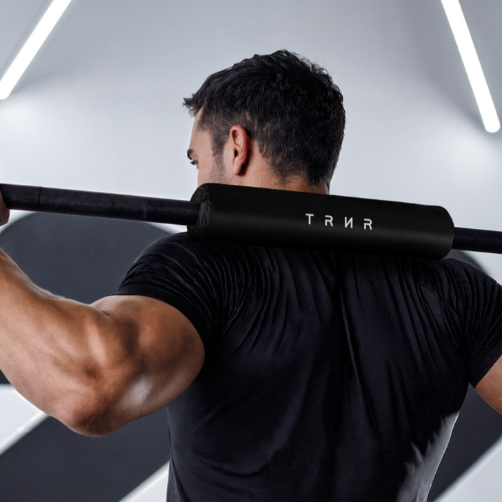 Person lifting a barbell with a neck pad featuring 'TRNR' branding in a gym setting