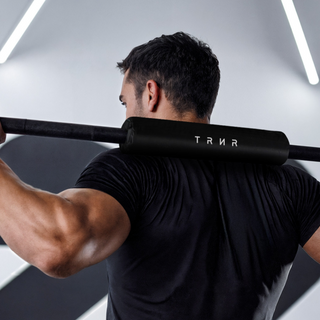Person lifting a barbell with a neck pad featuring 'TRNR' branding in a gym setting